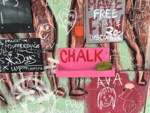 Colourful graphitti wall with messages involving discounts, Free Palestine, doodles. It has a prominent pink shelf in centre that reads CHALK. On it is a green sponge, but no chalk.