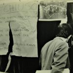 Black and white photo of back of man walking past a ripped handwritten sign which reads, "CHIUSO. Sotto questi condizioni non apriamo..." A photograph of riot police holding battons hangs next to it.