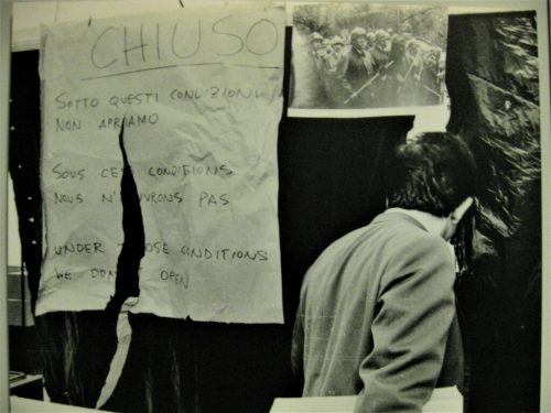 Black and white photo of back of man walking past a ripped handwritten sign which reads, "CHIUSO. Sotto questi condizioni non apriamo..." A photograph of riot police holding battons hangs next to it.