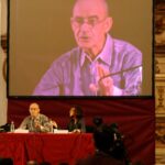Photograph of Jean Luc Nancy speaking in a grand venue and projected on a screen behind