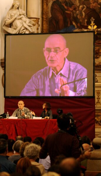 Photograph of Jean Luc Nancy speaking in a grand venue and projected on a screen behind
