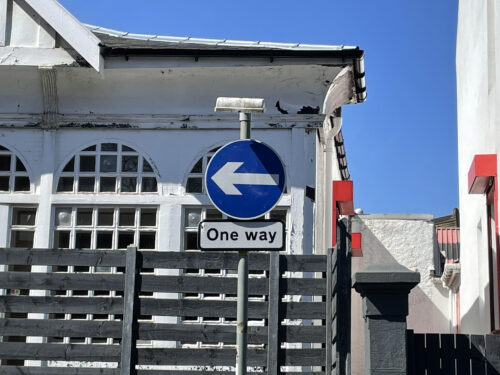 Blue one way street sign pointing left with white building behind