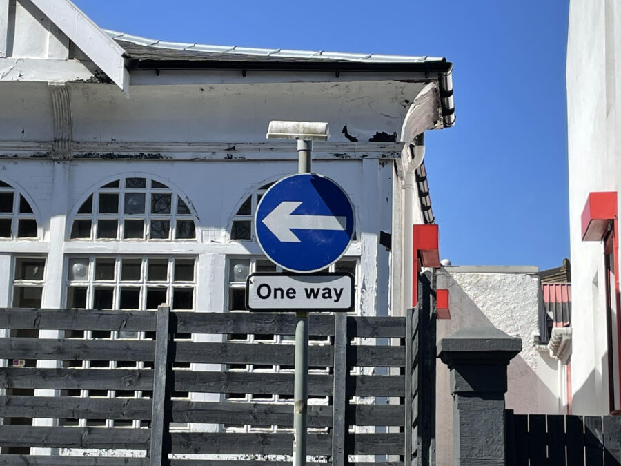 Blue one way street sign pointing left with white building behind